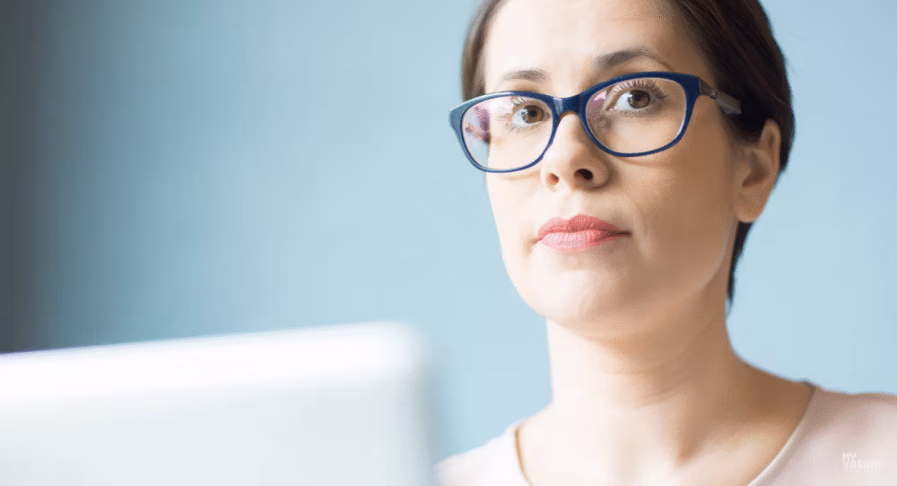 Woman in blue-framed glasses looking thoughtfully past an open laptop, seated against a pale blue-green wall